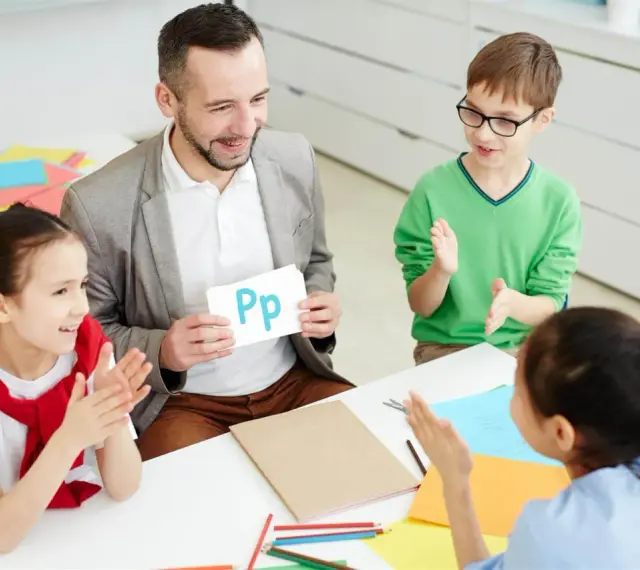 Nauczyciel pokazuje literę "Pp" podczas zebrania z rodzicami. Dzieci klaszczą, ucząc się alfabetu.