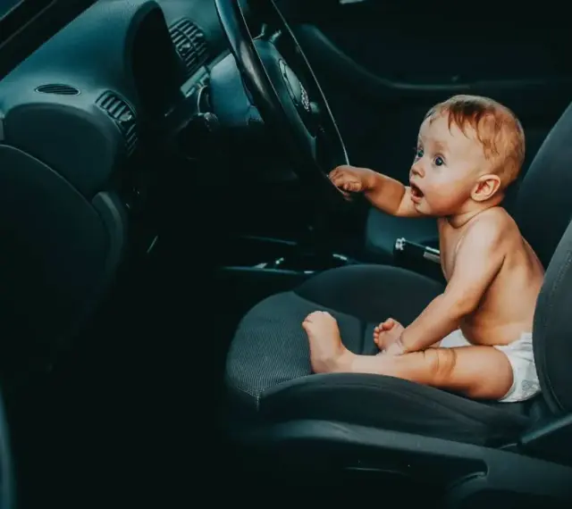 Maluch w pieluszce siedzi za kierownicą, jakby zastanawiał się, od kiedy dziecko może jeździć z przodu.