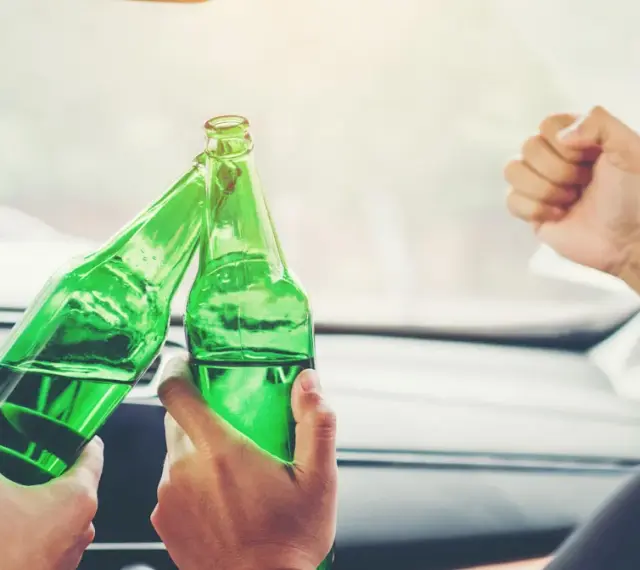 Dwie osoby w samochodzie wznoszą toast zielonymi butelkami. To nie jest dopuszczalna ilość alkoholu podczas jazdy.