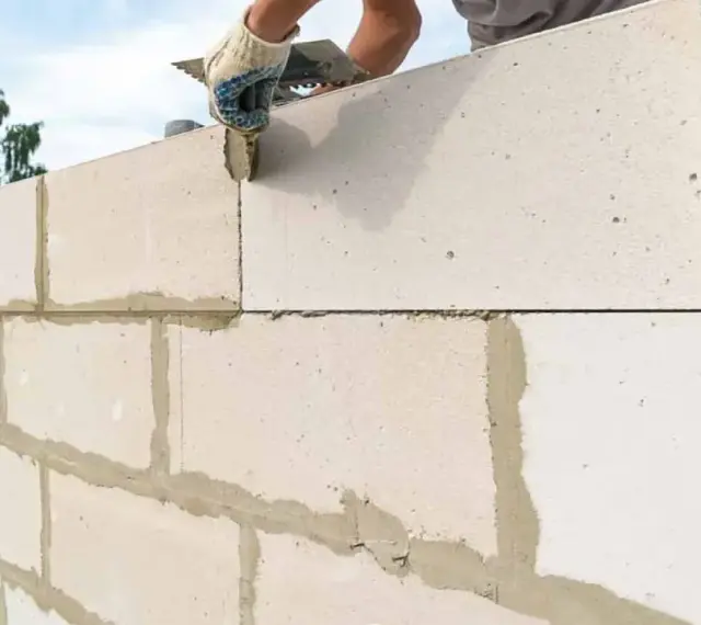Budowanie ściany z bloczków betonowych. Ręka w rękawicy nakłada zaprawę na kolejny element.