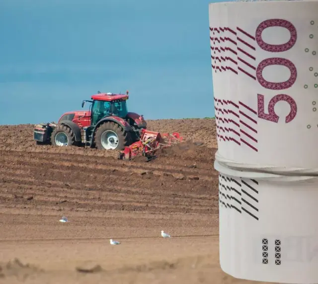 Czerwony traktor orze pole, a na pierwszym planie zwinięte banknoty 500 zł. Zastanawiasz się, ile kosztuje hektar ziemi budowlanej?