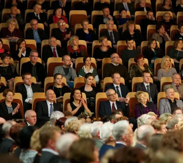 Elegancko ubrani ludzie siedzą w filharmonii. Panie w sukienkach, panowie w garniturach. To inspiracja, jak się ubrać do filharmonii.