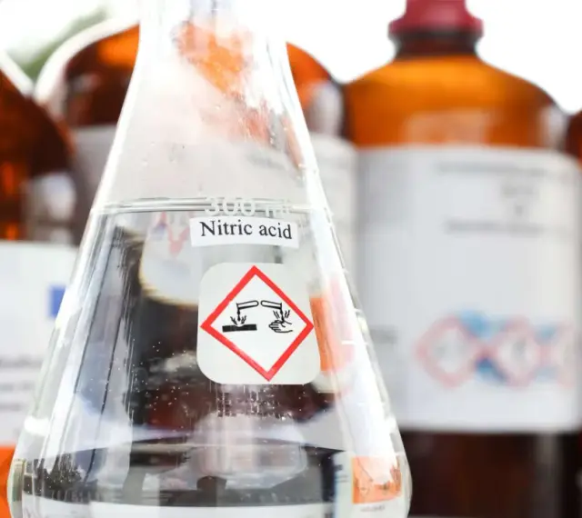 Ein Becherglas mit Salpetersäure (HNO3) und einem Gefahrensymbol. Im Hintergrund stehen weitere braune Glasflaschen mit Chemikalien.