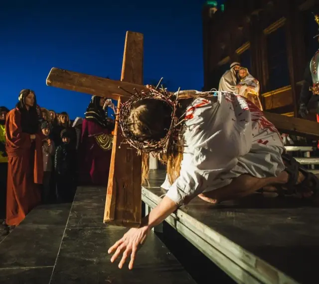 Scena drogi krzyżowej: Jezus w cierniowej koronie, z krwią na szacie, upada pod ciężarem krzyża. Tłum widzów obserwuje to poruszające przedstawienie.