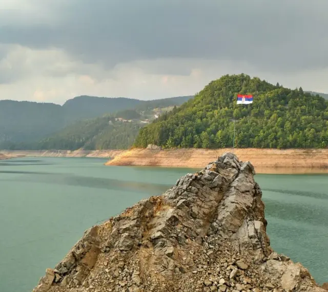 Serbska flaga na skale nad jeziorem. Krajobraz zachęca do podróży, a pytanie "czy Serbia jest bezpieczna dla turystów" wydaje się nie mieć znaczenia w obliczu tego spokoju.