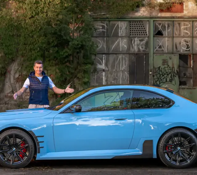 Mężczyzna prezentuje błękitne BMW M2 G87 przed zniszczonym budynkiem.