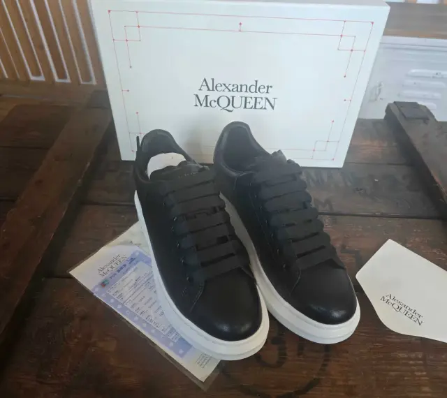 Czarne sneakersy Alexander McQueen na grubej, białej podeszwie, obok pudełka z logo marki.