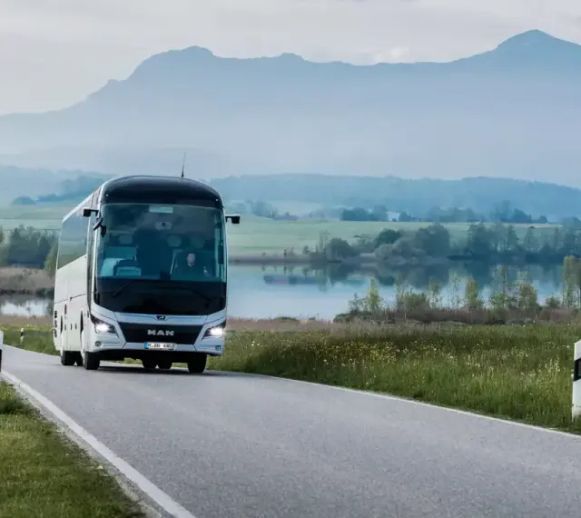 Biały **bus** marki **MAN** jedzie krętą drogą nad jeziorem, w tle góry.