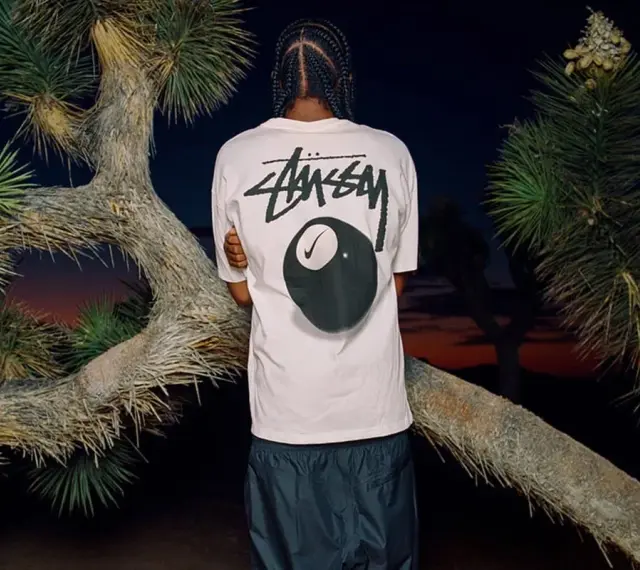 Mężczyzna w koszulce Stussy z grafiką kuli bilardowej 8 i logo Nike. Stussy to kultowa marka streetwearowa.