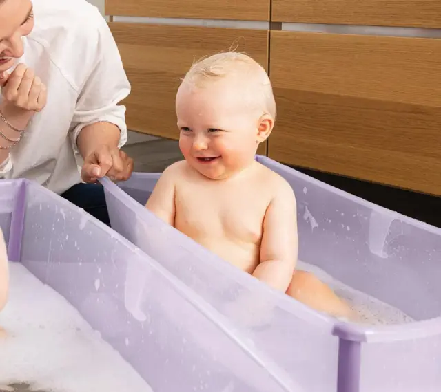 Uśmiechnięty bobas w fioletowym krzesełku do kąpieli, mama patrzy z czułością.