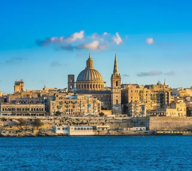 Malta, wyspa do kogo należy? Widok na Vallettę z kopułą i wieżą kościelną, otoczoną zabytkową architekturą, nad błękitnym morzem.