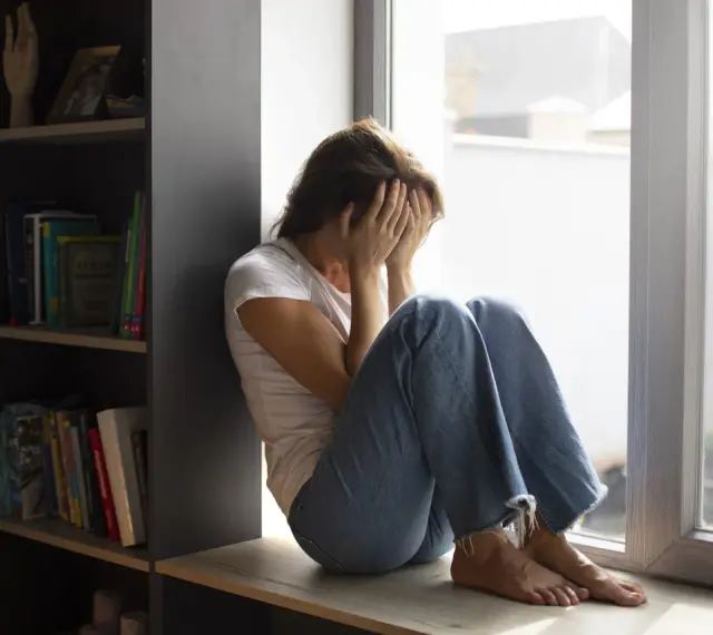 Kobieta siedzi przy oknie, zakrywając twarz dłońmi. Wokół panuje nastrój melancholii, odzwierciedlający dystymię.