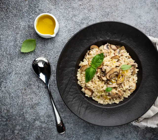 Kremowe risotto z grzybami na czarnym talerzu, obok łyżka i oliwa.