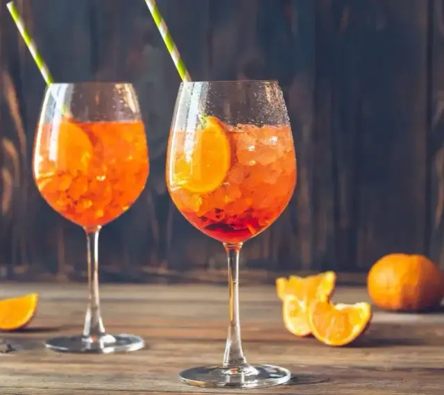 Dwa orzeźwiające drinki z aperolem, ozdobione plastrami pomarańczy i słomkami, na drewnianym blacie.