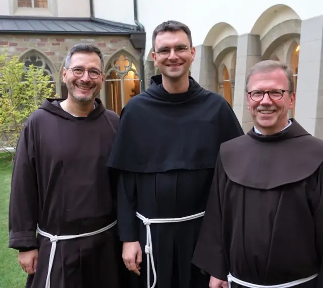 Drei Franziskaner Minoriten in braunen und schwarzen Kutten lächeln in die Kamera vor einer Klosterfassade.