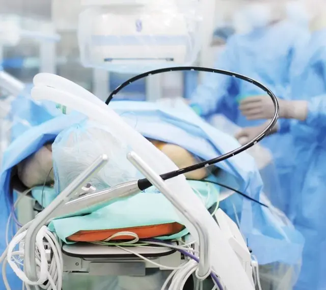Pacjent na stole operacyjnym, przygotowany do zabiegu PCI kardiologia. W tle personel medyczny w niebieskich fartuchach.