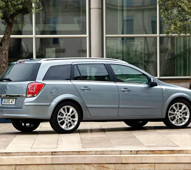 Srebrny Opel Astra H kombi zaparkowany przed nowoczesnym budynkiem z drzewami.