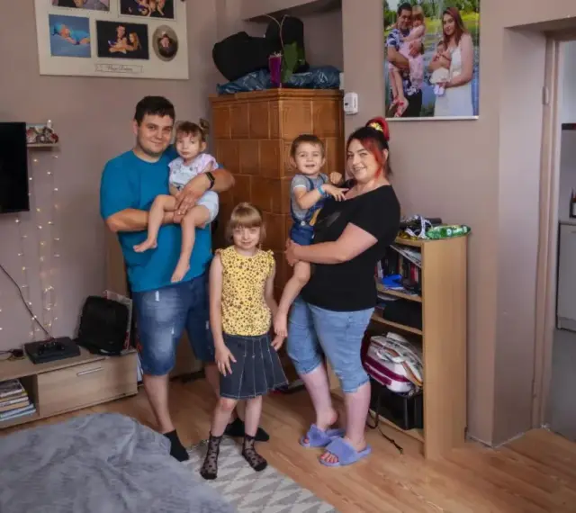 Rodzina z trójką dzieci pozuje do zdjęcia w salonie. W tle widać zdjęcia, telewizor i ozdobne lampki. Ich życie na kredyt nie przeszkadza im w cieszeniu się chwilą.