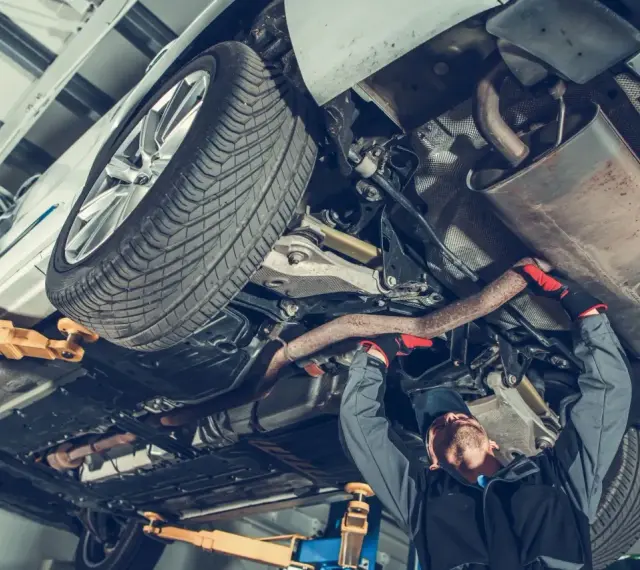 Mechanik pracuje pod samochodem, naprawiając budowę układu kierowniczego. Widać podniesione auto, koło i elementy wydechu.