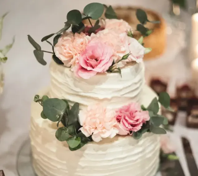 Elegancki tort weselny z kremową dekoracją i pastelowymi kwiatami, ozdobiony gałązkami eukaliptusa.