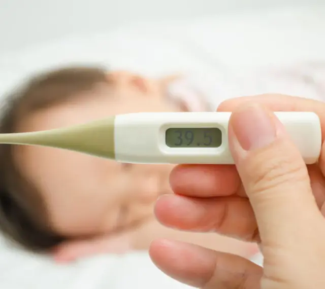 Cyfrowy termometr pokazuje 39.5°C. W tle widać śpiące dziecko. Zastanawiasz się, ile czasu mierzyć temperaturę pod pachą?