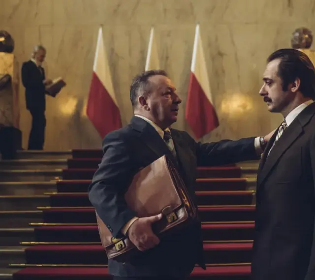 Hubert Niedziela, w garniturze, trzyma teczkę, rozmawiając z mężczyzną na schodach przed polskimi flagami.