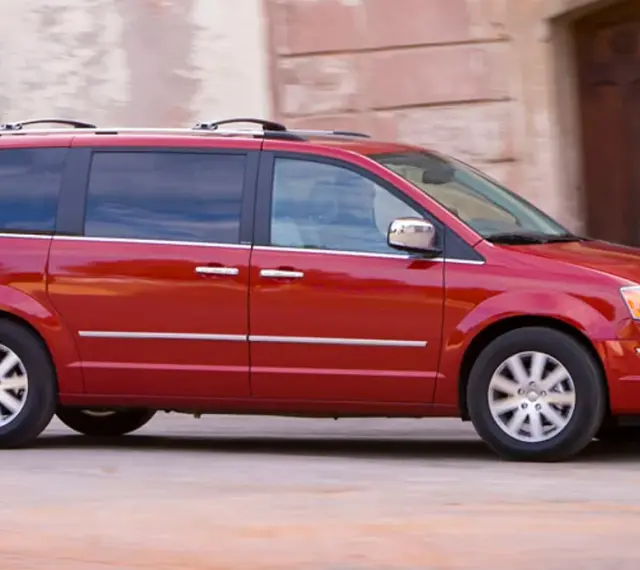 Czerwony Chrysler Grand Voyager w ruchu, z przyciemnianymi szybami i relingami dachowymi, przemierza ulicę obok kamiennej fasady.