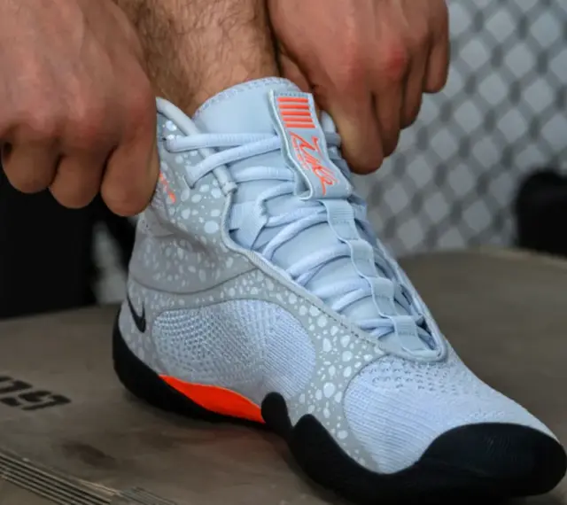 Ręce zapinają jasnoniebieskie buty sportowe z pomarańczowymi akcentami, przypominające styl Batistini.