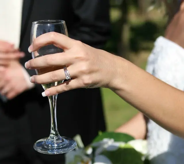 Dłoń panny młodej z pierścionkiem trzyma kieliszek szampana. W tle pan młody. 38 rocznica ślubu nazwa.