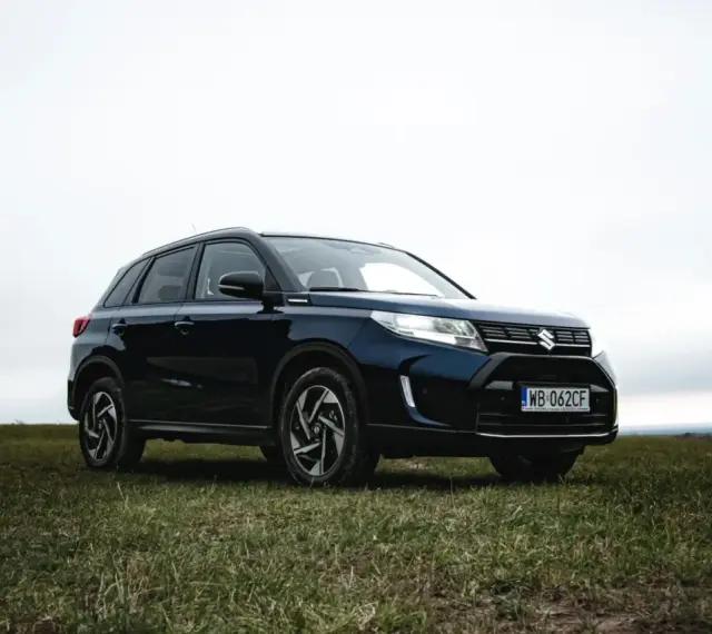 Ciemnoniebieski Suzuki Vitara Hybrid na tle zachmurzonego nieba, zaparkowany na trawiastym wzgórzu.