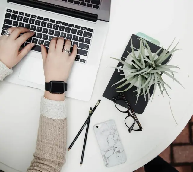 Kobieta pracuje na laptopie, obok leżą okulary, telefon i roślina. To idealne zdjęcia w chmurze do inspiracji.