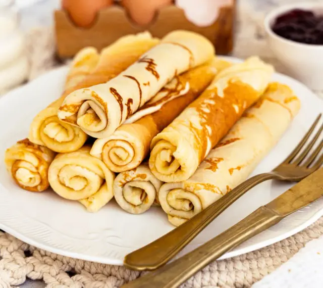 Ein Teller mit goldbraunen Quark Pfannkuchen, aufgerollt und gestapelt. Daneben liegen ein goldenes Messer und eine Gabel. Im Hintergrund eine Schale Marmelade.