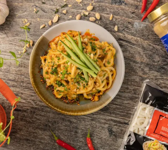 Makaron udon w sosie orzechowym, udekorowany ogórkiem i szczypiorkiem. Obok opakowanie makaronu udon mrożony czy suszony.