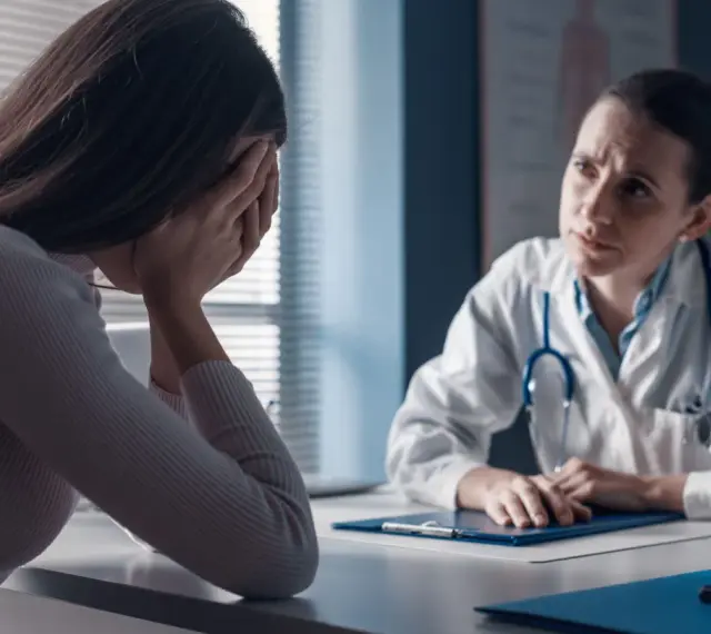 Kobieta w gabinecie lekarskim, ze smutkiem ukrytym w dłoniach, słucha diagnozy. Lekarka z troską pochyla się nad dokumentami, być może omawiając poronienie chybione.