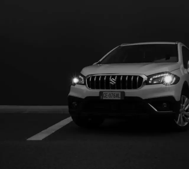 Białe Suzuki SX4 na ciemnym tle, z włączonymi światłami.