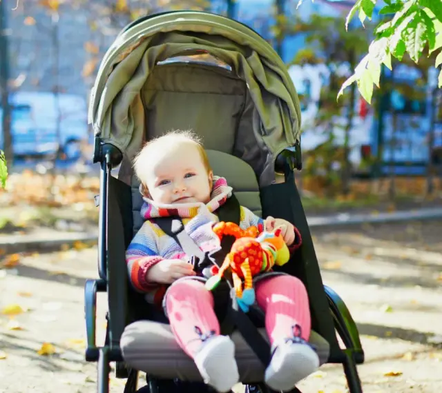 Uśmiechnięte dziecko w kolorowym sweterku i różowych skarpetkach siedzi w spacerówce, trzymając zabawkę. Czas na spacer!