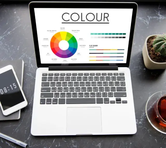Na ekranie laptopa koło barw i palety kolorów, pomocne przy wyborze między rgb czy cmyk do druku. Obok telefon, notes i kaktus.