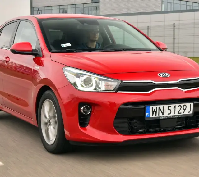 Czerwony Kia Rio w ruchu. Kierowca w okularach za kierownicą. Wiele pozytywnych kia rio opinie o tym modelu.