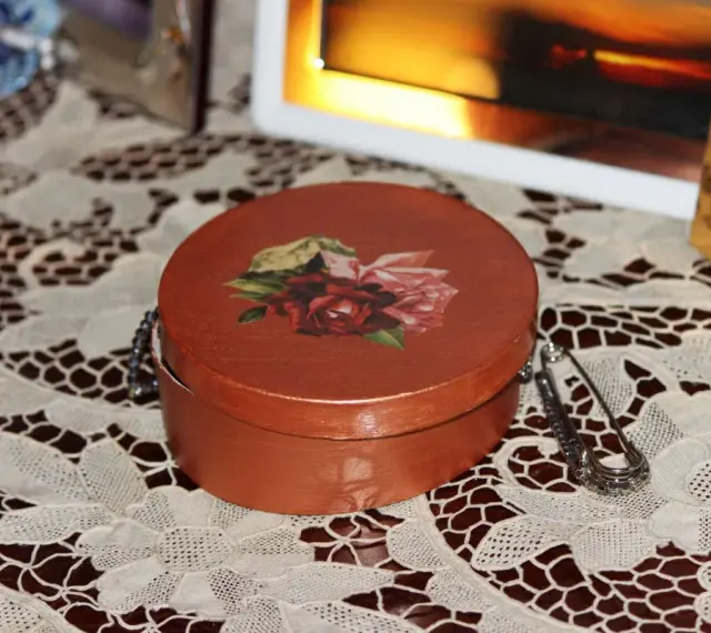 Okrągłe pudełko ozdobione techniką decoupage z motywem róż. Obok broszka i ramka ze zdjęciem.