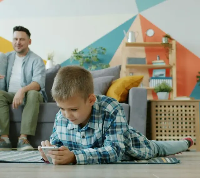 Chłopiec gra na telefonie, podczas gdy rodzice siedzą na kanapie. Ciekawe, od ilu lat jest WhatsApp i czy oni też go używają?
