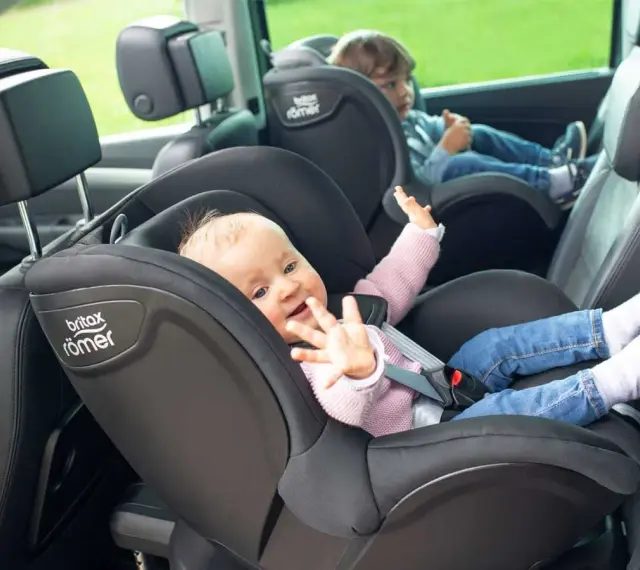 Uśmiechnięte niemowlę w foteliku Britax Römer, obok starsze dziecko. Od kiedy dziecko może jeździć z przodu? To pytanie nurtuje wielu rodziców.