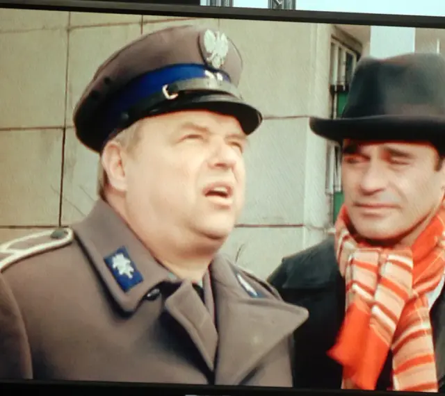 Dwóch mężczyzn w kapeluszach, jeden w mundurze, drugi w szaliku. Brunet wieczorową porą, w czarnym kapeluszu i czerwonym szaliku, rozmawia z policjantem.