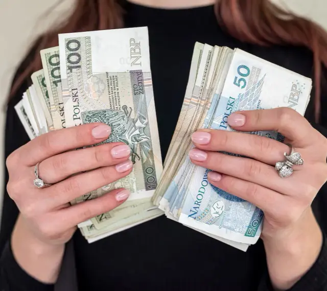 Dłonie z paznokciami w kolorze nude trzymają stosy banknotów 100 i 50 zł. To idealna jubileuszówka na prezent!