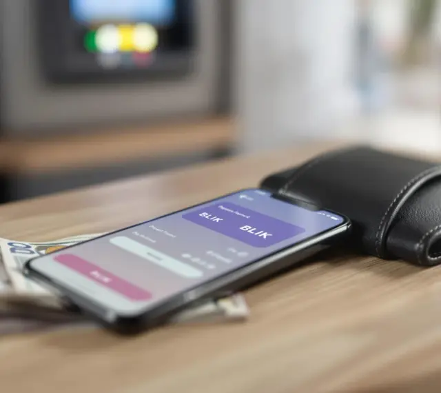 Smartfon z aplikacją BLIK, obok portfel i banknoty na drewnianym stole.