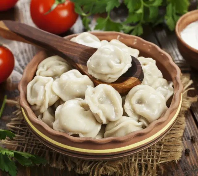 Pielmieni co to? Pyszne, gotowane pierożki w glinianej misce, z drewnianą łyżką, pomidorami i śmietaną w tle.