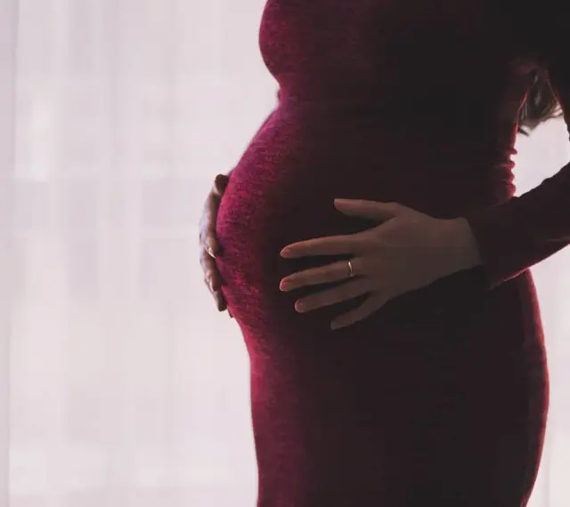 Kobieta w ciąży, z dłońmi na brzuchu, w ciemnoczerwonej sukience. Myśli o zasiłku macierzyńskim po ustaniu zatrudnienia.