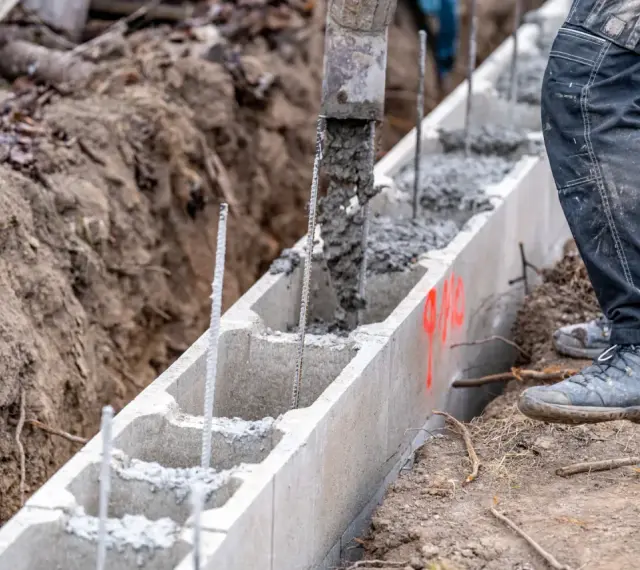 Beton wylewany do szalunku z bloczków betonowych, zbrojenie wystaje. Budowa fundamentów w toku.