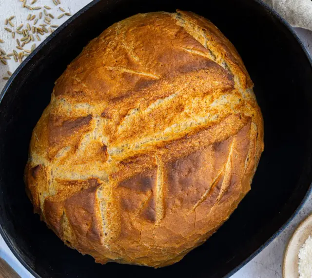 Ein goldbraunes Brot mit knuspriger Kruste liegt in einem Gusseisentopf. Dieses Brot kommt nicht aus dem Topf, sondern wurde darin gebacken.