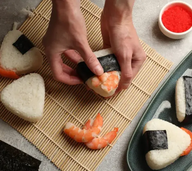 Ręce formują onigiri z krewetkami, tworząc pyszne japońskie jedzenie. Obok miska ryżu, nori i talerz gotowych przysmaków.