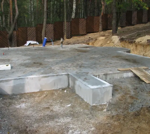 Solidna płyta fundamentowa, gotowa na budowę domu. Wokół las i drewniany płot.
